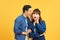 Portrait of asian man whispering secret or interesting gossip to excited woman in her ear isolated over yellow background