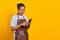 Portrait of Asian young man wearing apron smiling broadly looking at incoming message on smartphone on yellow background