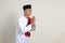 Portrait of attractive Asian muslim man in white shirt with skullcap showing apologize and welcome hand gesture. Isolated image on