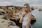 Portrait of attractive blonde woman with silence gesture posing on rocky beach with beauty ocean landscape.