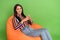 Portrait of attractive cheerful curious brunet girl sitting in bag chair pondering isolated over bright green color