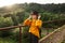 Portrait of an attractive female hiker in orange sweatshirt and hat standing on a bridge in the mountains and looking away with a