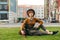 Portrait of attractive girl walking with dog, sitting on lawn, stroking puppy and posing at camera against cityscape background.