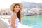 Portrait of attractive smiling latin woman with hat and summer dress looking at camera with Cefalu village on the background