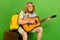 Portrait of attractive trendy cheerful guy hippie playing guitar sitting on valise isolated over bright green color