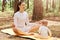 Portrait of attractive woman sitting in lotus pose and meditating while her infant daughter or son playing on karemat near mother