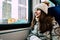 portrait of attractive young brunette woman wearing beige warm knitted hat with fur pompom and scarf. Girl looking at sityscape