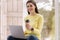 Portrait of attractive young woman using laptop in front of big window. Brunette woman in yellow sweatshirt working at
