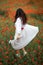 Portrait from back of young female with beautiful long hair and long white dress dancing on poppies field at summer