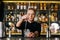 Portrait of bartender male stirring ice cubes in glass using spoon standing on bar counter, on background of shelves