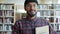 Portrait of bearded African American guy in hat holding books in library smiling