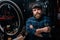 Portrait of bearded cycling mechanic male holding wrench in hand standing by bicycle in repair workshop with dark