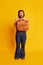 Portrait of bearded hairy man, shy scientist nerd in flared jeans standing with briefcase over yellow background. Retro
