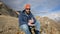 Portrait of a bearded traveler in sunglasses and a cap sits on a rock against the backdrop of mountains. Laughing and