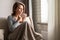 Portrait of beautiful Ñaucasian young woman sitting on sofa covered blanket, drinking hot coffee from cup and looking at window.