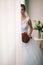 Portrait of beautiful bride in white silk dressing gown with curly hairstyle and long veil standing near window in bedroom