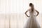 Portrait of beautiful bride in white silk dressing gown with curly hairstyle and long veil standing near window in bedroom, copy s