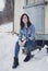 Portrait of beautiful fashion model posing to camera and sitting on the stone in front the caravan in winter time. Full body shot