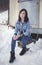 Portrait of beautiful fashion model posing to camera and sitting on the stone in front the caravan in winter time. Full body shot