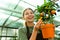 Portrait of beautiful florist woman 20s holding orange tree while working in greenhouse