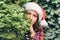 Portrait of a beautiful girl with a dreaming, piercing look in a santa claus red hat looks away through green branches of juniper.