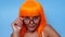 Portrait of beautiful happy Asian woman in bright orange wig and glasses posing over blue