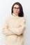 portrait of a beautiful, happy, neat woman in a beige turtleneck sweater and black-rimmed glasses, standing on a light