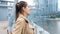 Portrait of beautiful smiling woman standing on bridge and looking on modern buildings and calm river