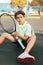 Portrait of a beautiful teenage boy with a tennis racket in his hands. A tennis player sits on the court and rests after