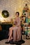 Portrait of beautiful woman in evening dress sitting with glass of champagne in decorated room with heap of christmas gifts