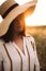 Portrait of beautiful woman in hat enjoying sunset golden light in summer meadow. Stylish rustic girl in linen dress relaxing in