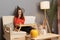 Portrait of beautiful woman wearing witch hat working on laptop in home interior and drinking wine, holding glass of alcohol and