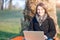 Portrait of a beautiful young brunette woman working on laptop sitting at the foot of a tree at the park. She smiles at the camera