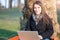 Portrait of a beautiful young brunette woman working on laptop sitting at the foot of a tree at the park. She smiles at the camera