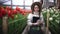 Portrait of beautiful young female smiling woman worker florist in apron and hat stands with tablet in greenhouse and