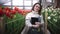 Portrait of beautiful young female smiling woman worker florist in apron stands with tablet in greenhouse and looks at