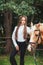 Portrait beautiful young girl in white shirt and black pants with beauty long hair next horse in forest. Fashionable elegance woma