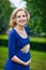 Portrait of beautiful young pregnant woman in blue dress with long blond curly hair holding her belly and smiling