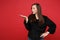 Portrait of bewildered young woman in black fur sweater looking, pointing hand aside isolated on bright red wall