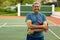 Portrait of biracial happy senior man with arms crossed holding balls and racket at tennis court