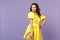 Portrait of blinking young woman in yellow dress holding summer hat, biting lips isolated on pastel violet wall