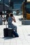 Portrait of blond girl with blue dress and phone waiting with suitcase in front of the train station