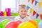 Portrait of blonde caucasian boy smiling at camera near birthday rainbow cake. Festive colorful background with balloons