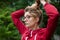 Portrait of blonde girl with hipster eyeglasses in dark red jacket making ponytail outdoors in the urban park