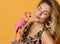 Portrait of blonde woman in sundress with tropical print holding pink flamingo statuette sending a kiss on yellow