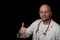 Portrait of a bold doctor with red stethoscope on dark background. Looking at the viewer. Thumbs up gesture with his right hand.