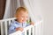 Portrait of bored baby standing in crib. Baby boy stand alone in crib. Sad little baby