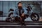 Portrait of a brutal bearded male dressed in a jeans jacket sitting on steps near his custom-made retro motorcycle