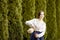 Portrait of Calm Standing Caucasian Girl in Rural Bright Dress Pouring Milk From Clay Jar in Front of Fir Trees