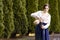 Portrait of Calm Standing Caucasian Girl in Rural Bright Dress Pouring Milk From Clay Jar in Front of Fir Trees Outdoors